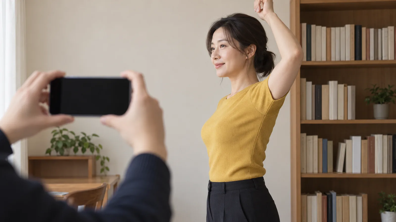 책장 옆에서 겨자색 반팔 니트를 입은 여성이 한쪽 팔을 들고, 앞쪽 두 손이 꺼진 화면의 휴대폰을 가로로 들어 그 모습을 찍는 장면