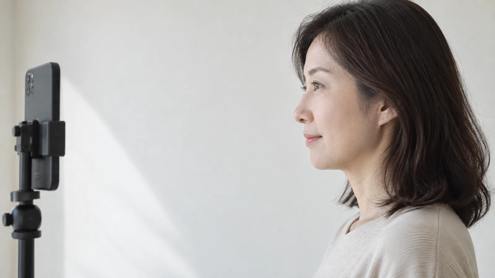 무늬 없는 밝은 벽 앞에 선 40대 여성의 측면 모습과, 화면 왼쪽 눈높이의 삼각대에 세운 휴대전화 뒷면이 보인다.